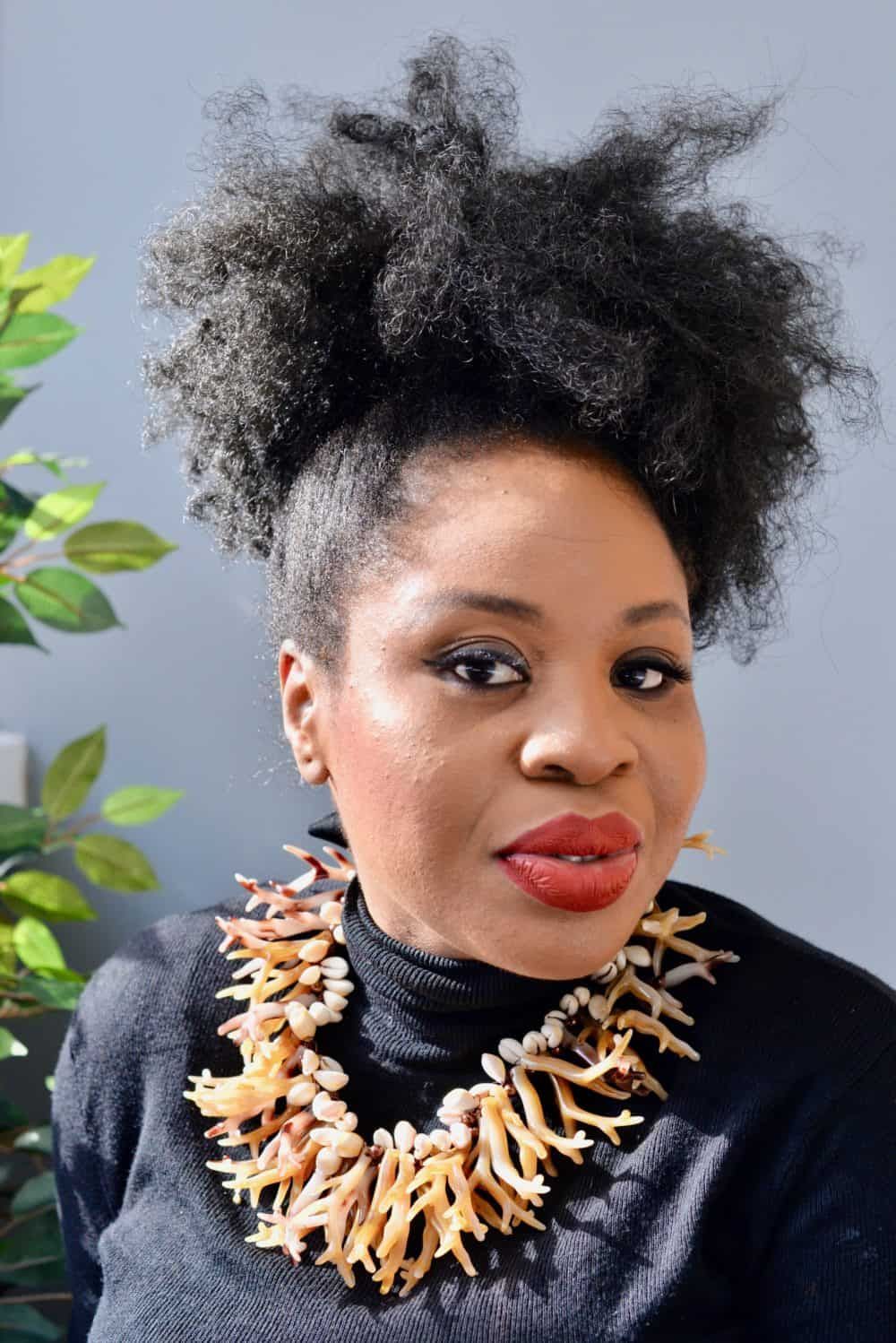 Portrait of Dr Claire Ogah, artist and founder of Overlay Couture, wearing a sculpted top-knot and coral statement necklace